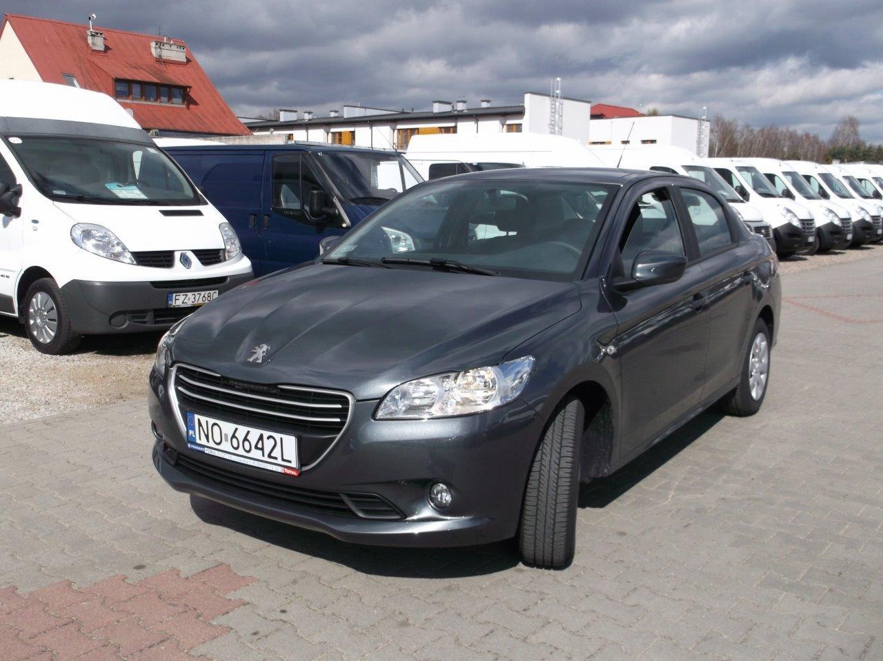 Szary Peugeot 301 na parkingu z rzędem białych busów w tle, widoczne tablice rejestracyjne.