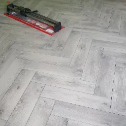 Ułożona w jodełkę podłoga z paneli imitujących jasne drewno, obok leży przecinarka do płytek i wiadro z akcesoriami.