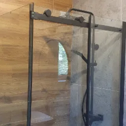 Nowoczesna kabina prysznicowa z czarnymi profilami i deszczownicą, wykończona płytkami imitującymi drewno i szarym kamieniem. Widoczny fragment siedziska.