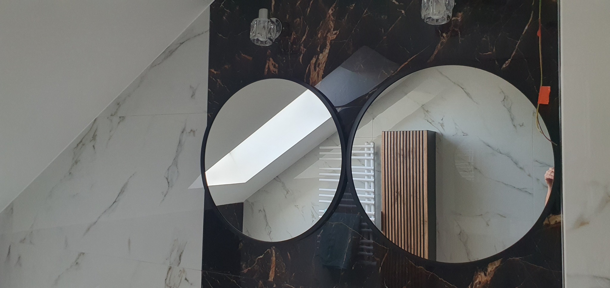 Wykończona łazienka na poddaszu z dwoma okrągłymi lustrami i marmurowymi płytkami. Widoczny grzejnik drabinkowy i szafka z lamelami.