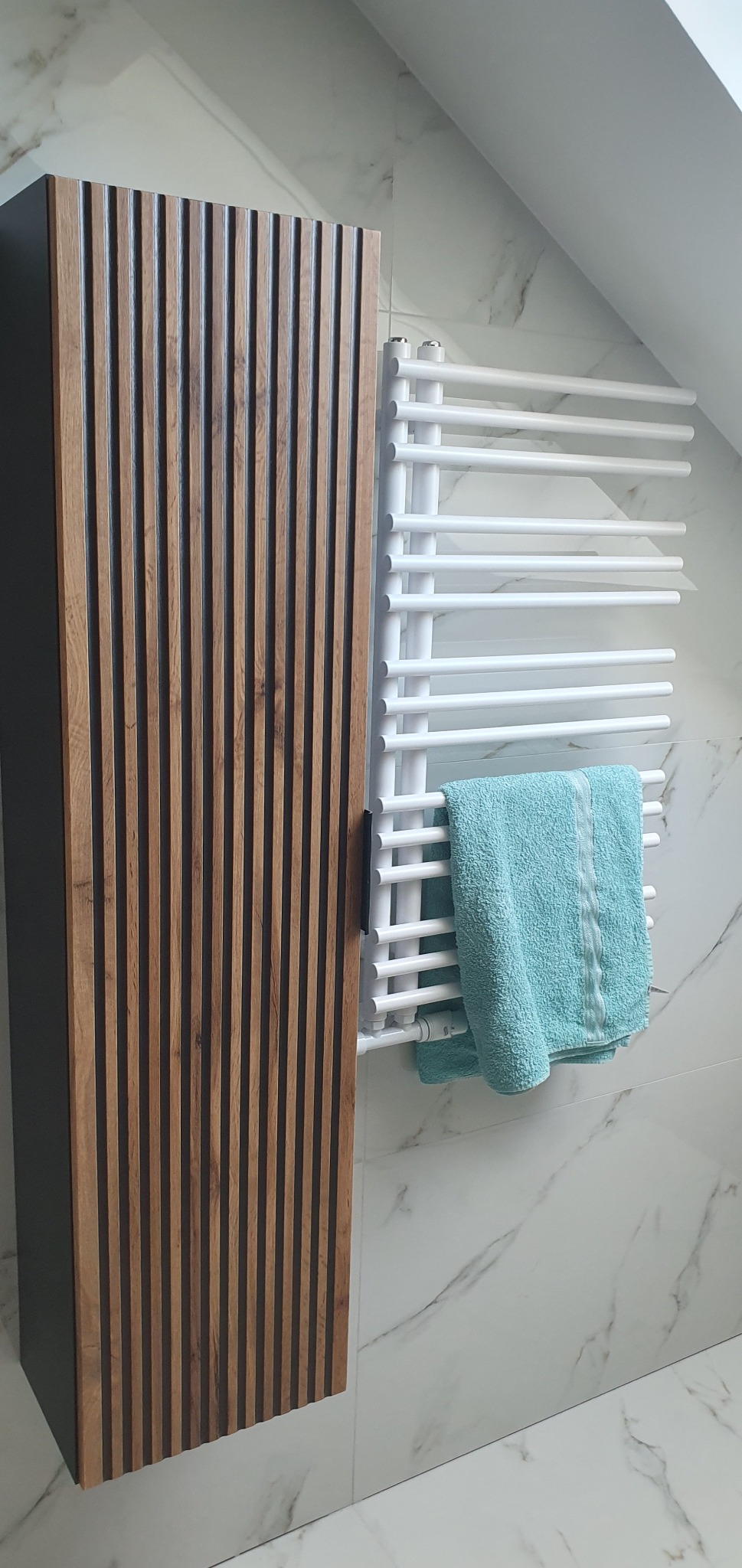 Biały grzejnik łazienkowy z turkusowym ręcznikiem, obok szafka z drewnianym frontem na tle marmurowych płytek w łazience na poddaszu.