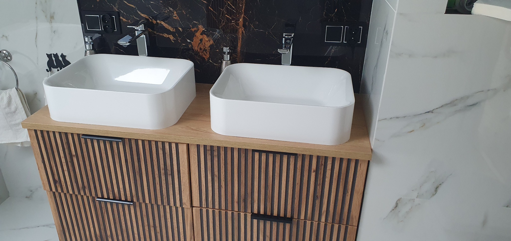 Nowoczesna łazienka: dwie białe, kwadratowe umywalki nablatowe na szafce z drewnianymi lamelami, marmurowe płytki na ścianie i czarne akcenty.