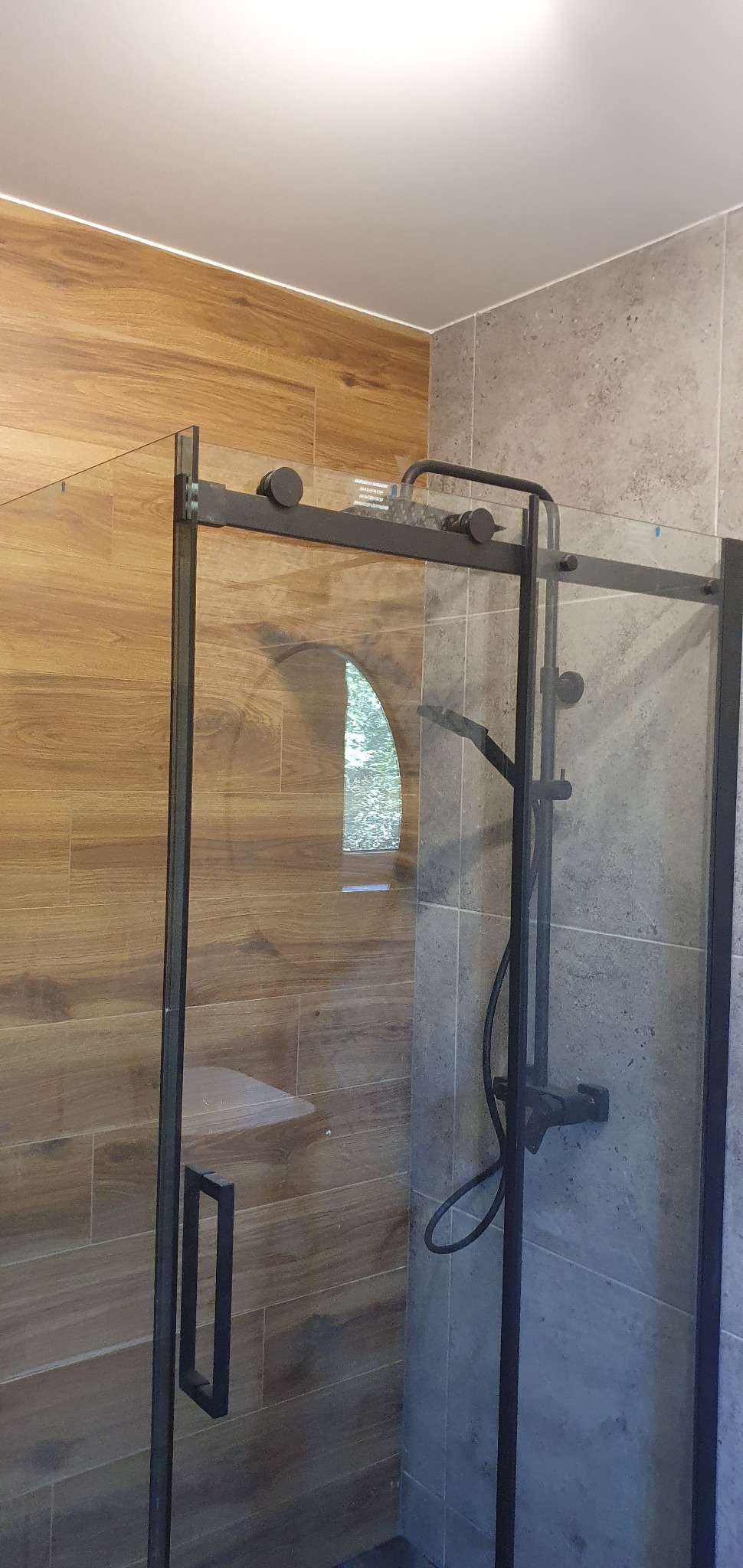 Nowoczesna kabina prysznicowa z czarnymi profilami i deszczownicą, wykończona płytkami imitującymi drewno i szarym kamieniem. Widoczny fragment siedziska.