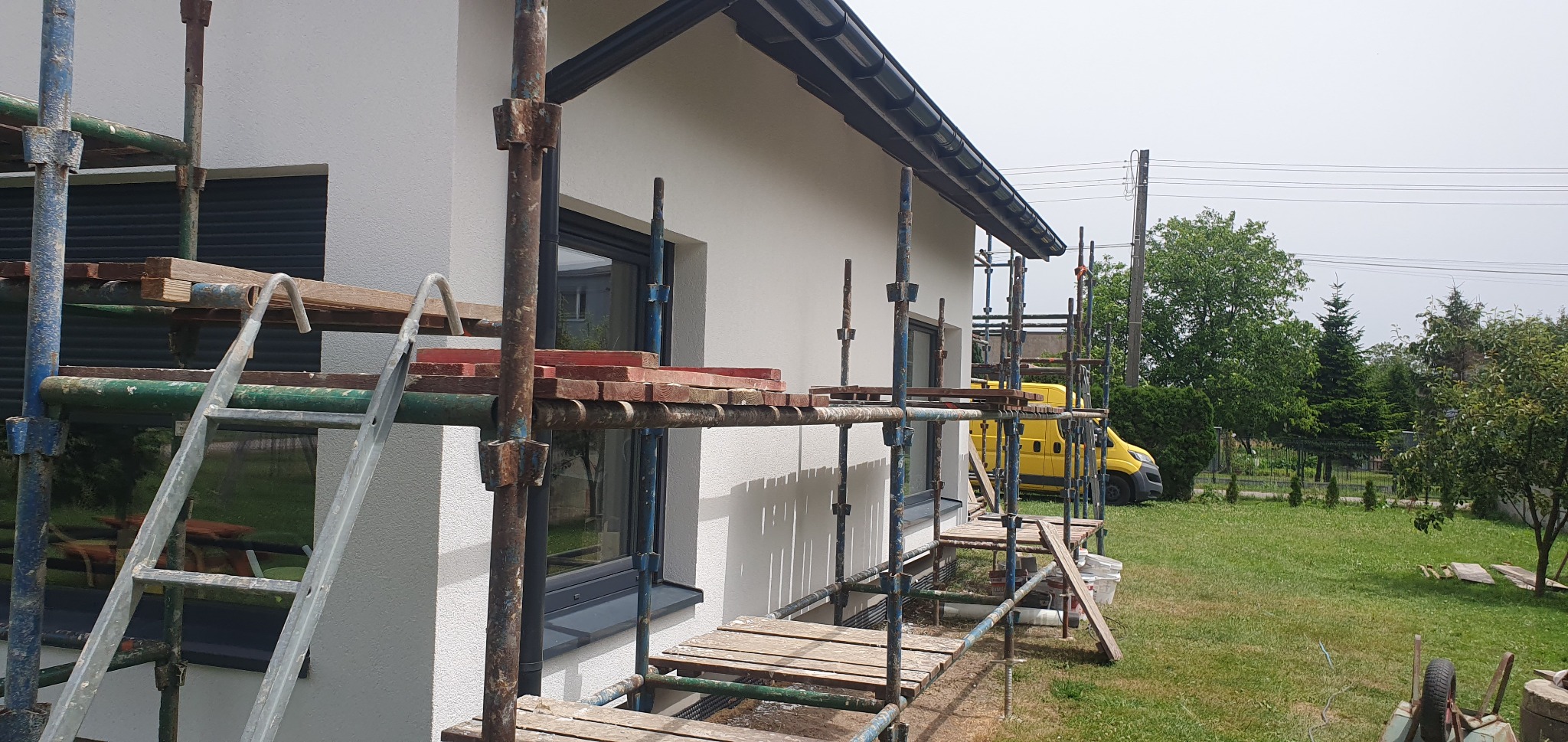 Biały dom jednorodzinny w trakcie prac elewacyjnych, widoczne rusztowania i drabina. W tle zieleń drzew i krzewów, oraz żółty samochód dostawczy.