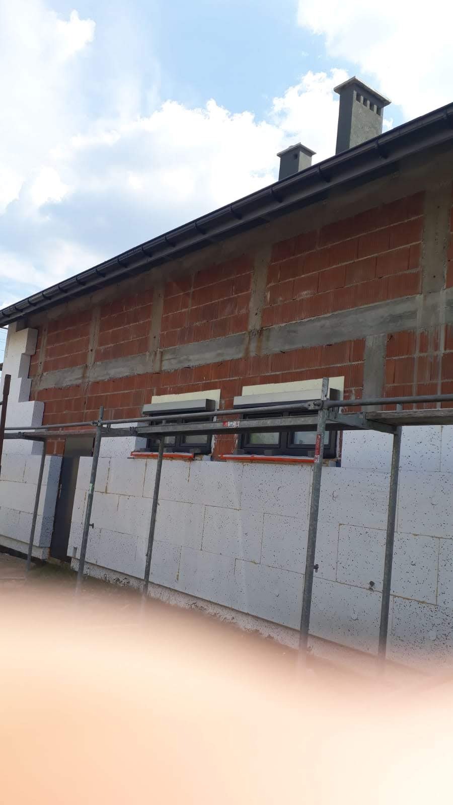 Budynek w trakcie ocieplania styropianem, widoczne rusztowania i okna. Częściowo ocieplona ściana z czerwonej cegły. Dwa kominy na dachu.