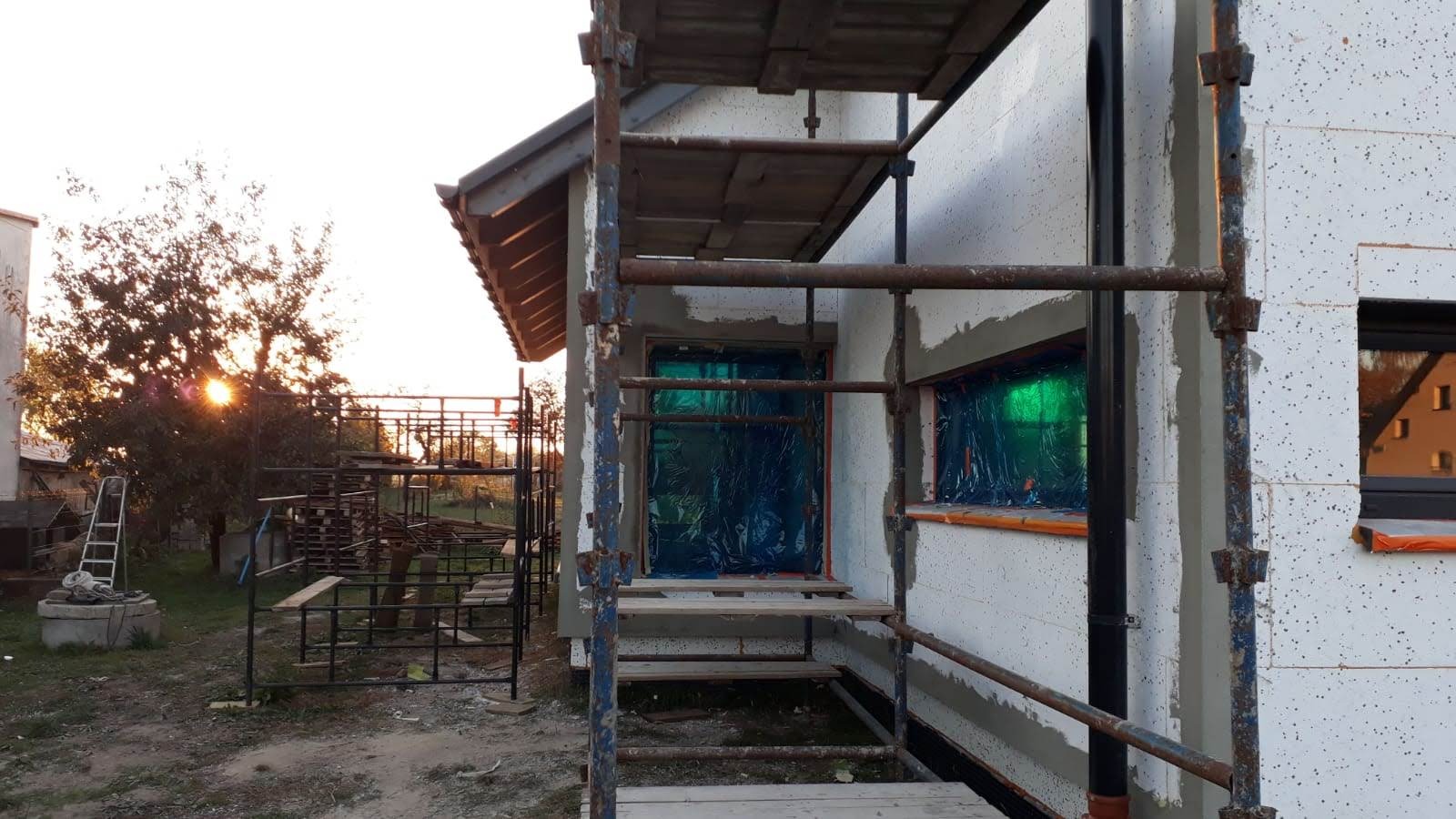 Ocieplanie elewacji budynku jednorodzinnego z użyciem rusztowania. Okna i drzwi zabezpieczone folią. Widoczne płyty styropianowe i warstwa kleju.