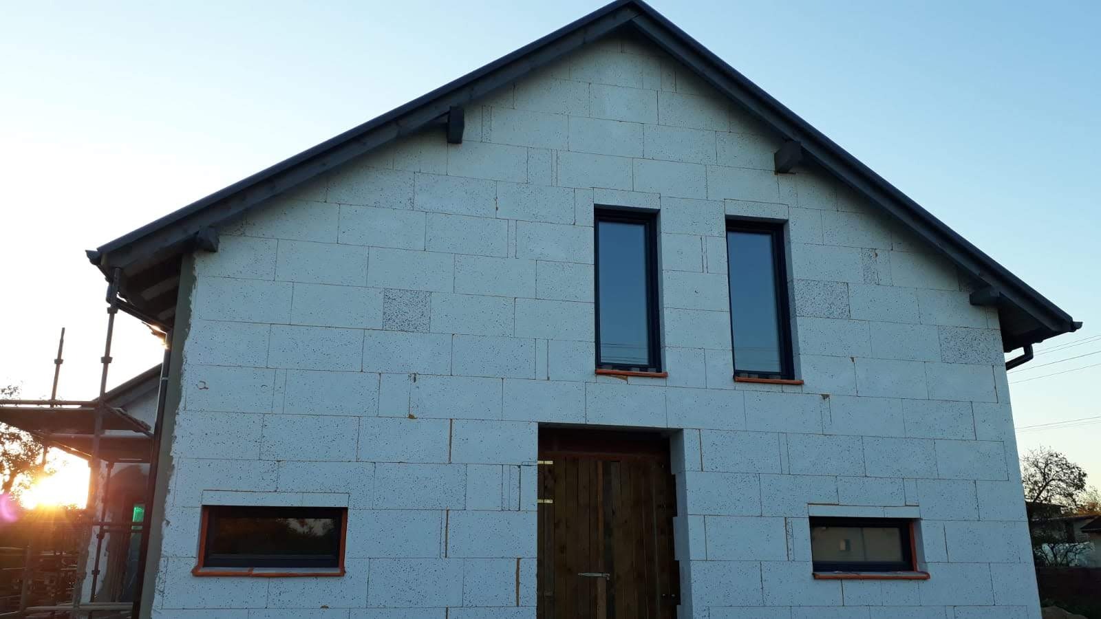 Surowy stan budynku z betonu komórkowego z zamontowanymi oknami i drzwiami wejściowymi. Widoczne rusztowania i elementy dachu. Ujęcie z dołu, wczesny etap budowy.