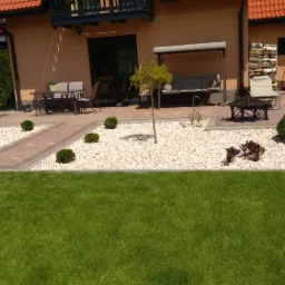 Ujęcie ogrodu z trawnikiem, ścieżką z kostki, rabatami z białym kamieniem, ozdobnymi krzewami i drzewami, z tarasem przy domu z meblami ogrodowymi i huśtawką.