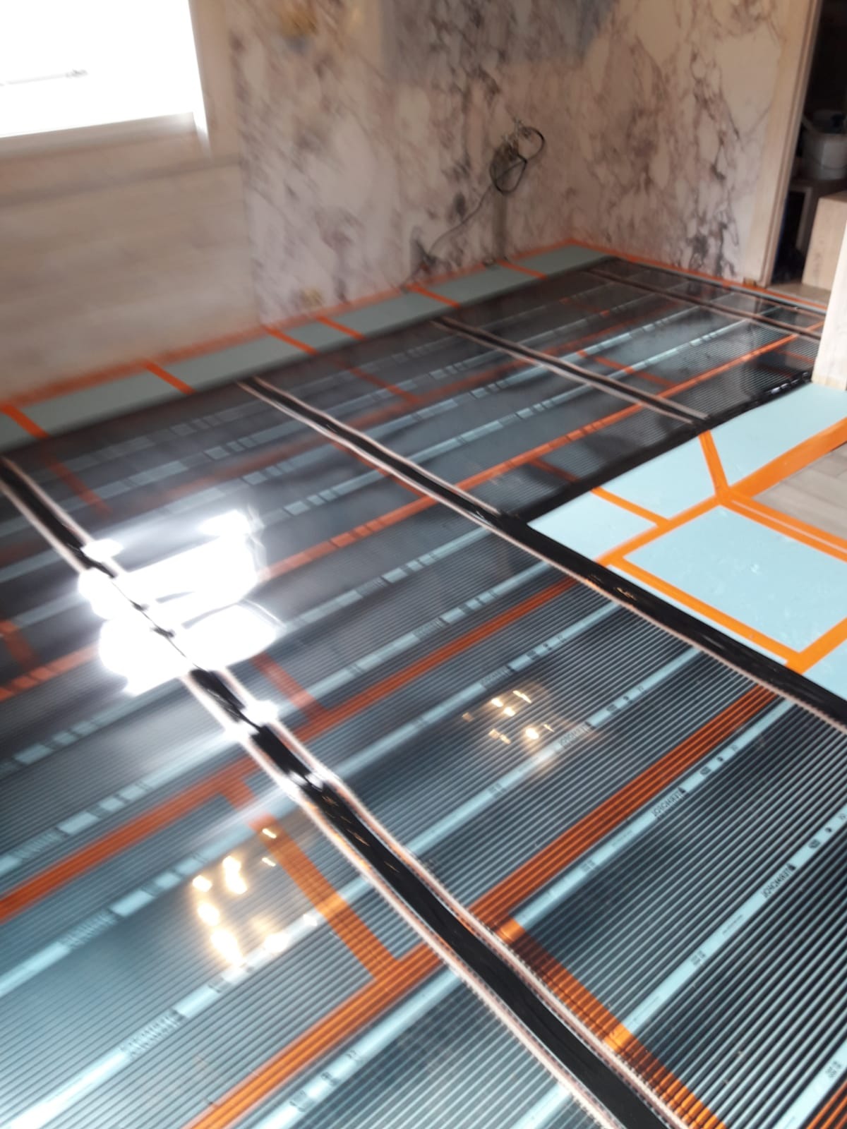 Montaż maty grzewczej na podłodze w Górowie Iławeckim, widoczne czarne pasy grzewcze ułożone na izolacji termicznej z pomarańczowymi elementami mocującymi.