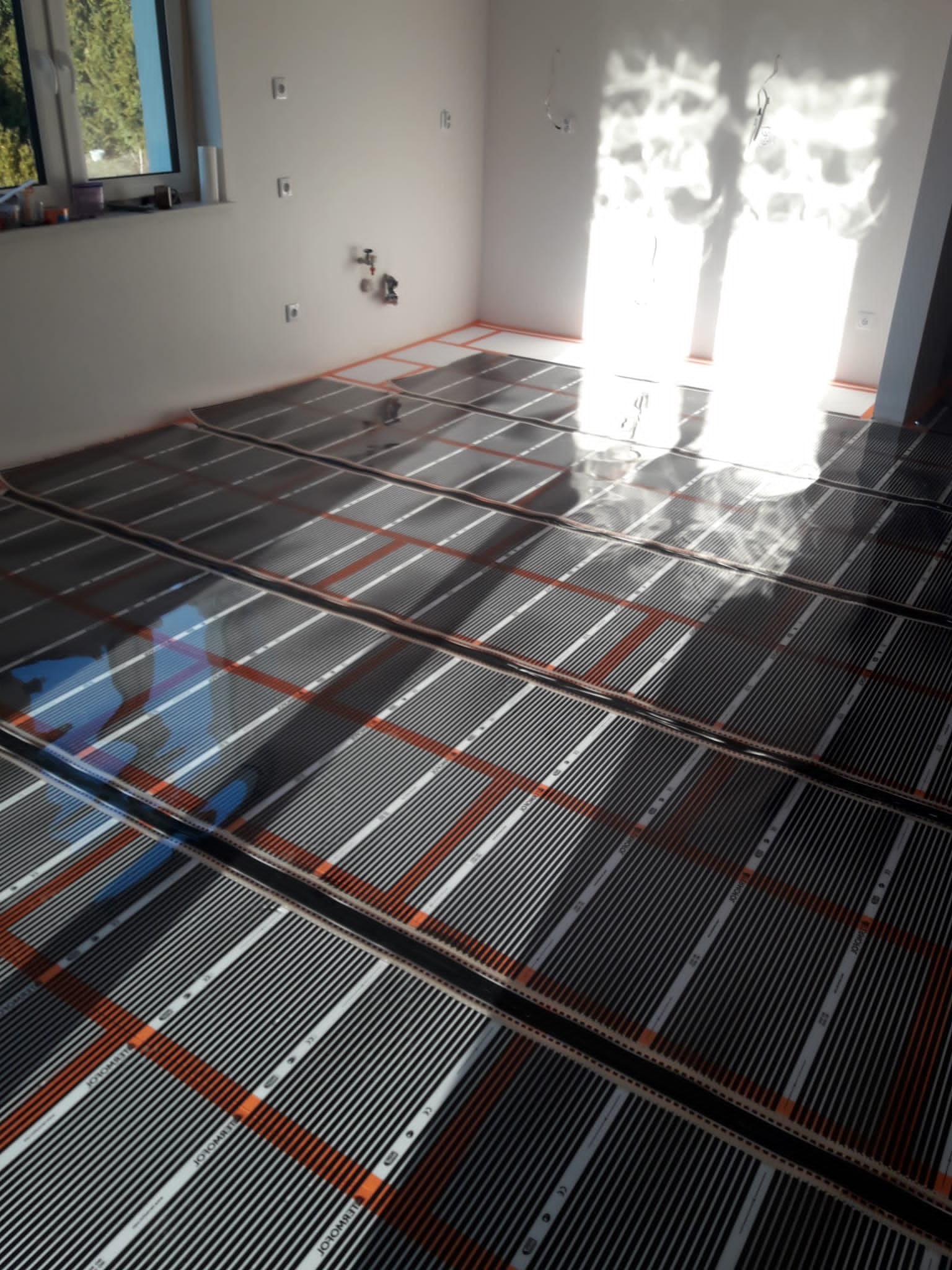 Montaż mat grzewczych pod ogrzewanie podłogowe w jasnym pomieszczeniu z oknem, widoczne kable i instalacja na ścianie.