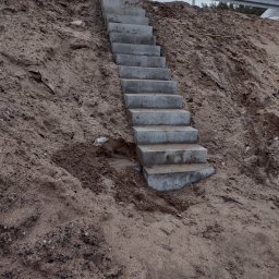 Przedsiębiorstwo Handlowo Usługowe JP Paulina Jastruszewska - Betonowe schody prowadzą w górę po zboczu ziemi, zabezpieczone metalową barierką. Widok na szary, pochmurny dzień. Wykonanie z surowego betonu.