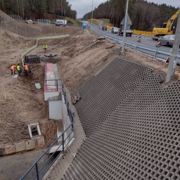 Przedsiębiorstwo Handlowo Usługowe JP Paulina Jastruszewska - Budowa drogi: robotnicy w kamizelkach przy betonowym korycie, w tle droga z barierami, koparka i las. Ułożona ażurowa kostka na zboczu.