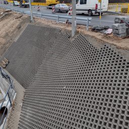 Przedsiębiorstwo Handlowo Usługowe JP Paulina Jastruszewska - Wzmocnienie skarpy przy drodze za pomocą ażurowych płyt betonowych. Widoczne elementy infrastruktury drogowej i fragment przepustu wodnego.