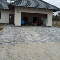 Ułożona kostka brukowa przed domem z otwartym garażem, widoczne prace wykończeniowe i przygotowanie terenu. Szara kostka w geometryczny wzór.