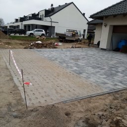 Przedsiębiorstwo Handlowo Usługowe JP Paulina Jastruszewska - Częściowo ułożona kostka brukowa na podjeździe, widoczne etapy prac. W tle domy jednorodzinne i samochód dostawczy. Taśma ostrzegawcza wyznacza obszar robót.