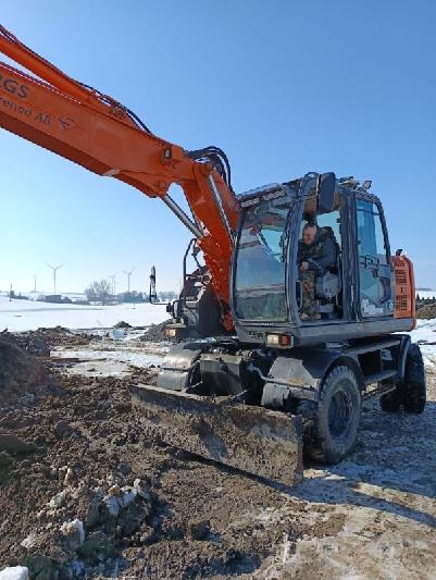 Pomarańczowa koparka kołowa z operatorem na placu budowy w zimowej scenerii. W tle widoczne turbiny wiatrowe i resztki śniegu.