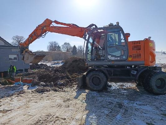 Pomarańczowa koparka kołowa w trakcie prac ziemnych na placu budowy, z widocznym operatorem w kabinie i pracownikiem w odblaskowej kamizelce w tle.