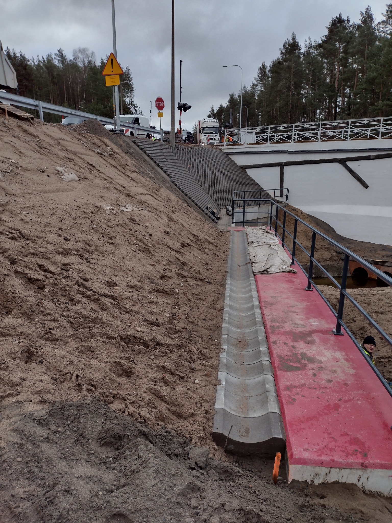 Skarpa z betonowymi schodami i korytem odwadniającym. Widoczny fragment mostu i znak drogowy. Prace ziemne w toku, teren przygotowany pod dalsze roboty.