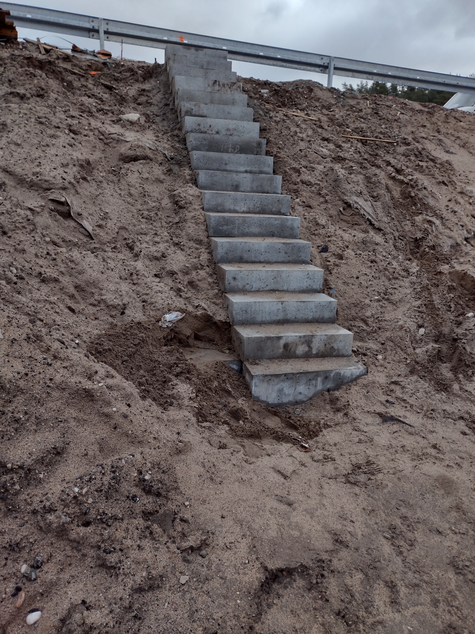Betonowe schody prowadzą w górę po zboczu ziemi, zabezpieczone metalową barierką. Widok na szary, pochmurny dzień. Wykonanie z surowego betonu.