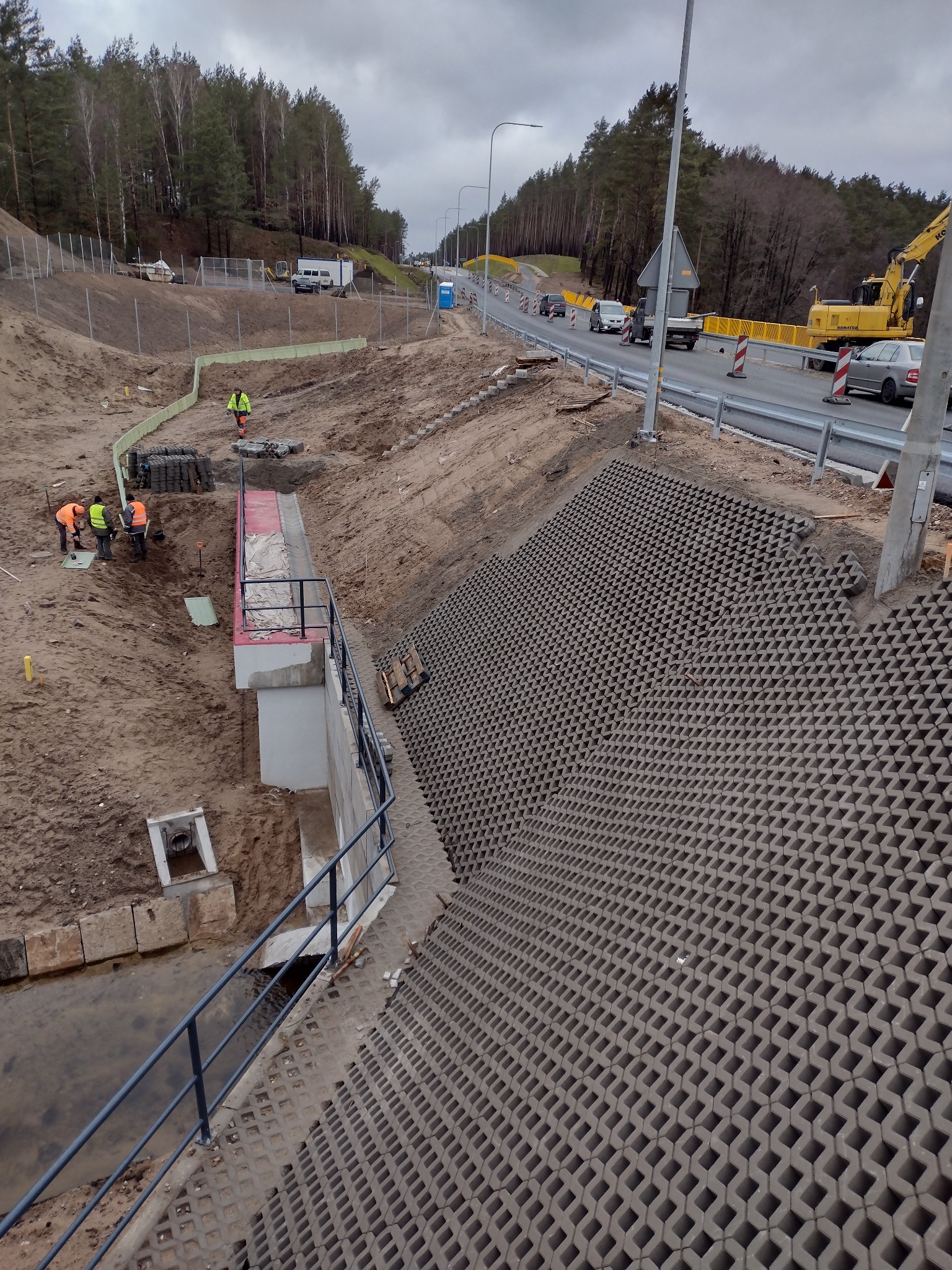 Budowa drogi: robotnicy w kamizelkach przy betonowym korycie, w tle droga z barierami, koparka i las. Ułożona ażurowa kostka na zboczu.