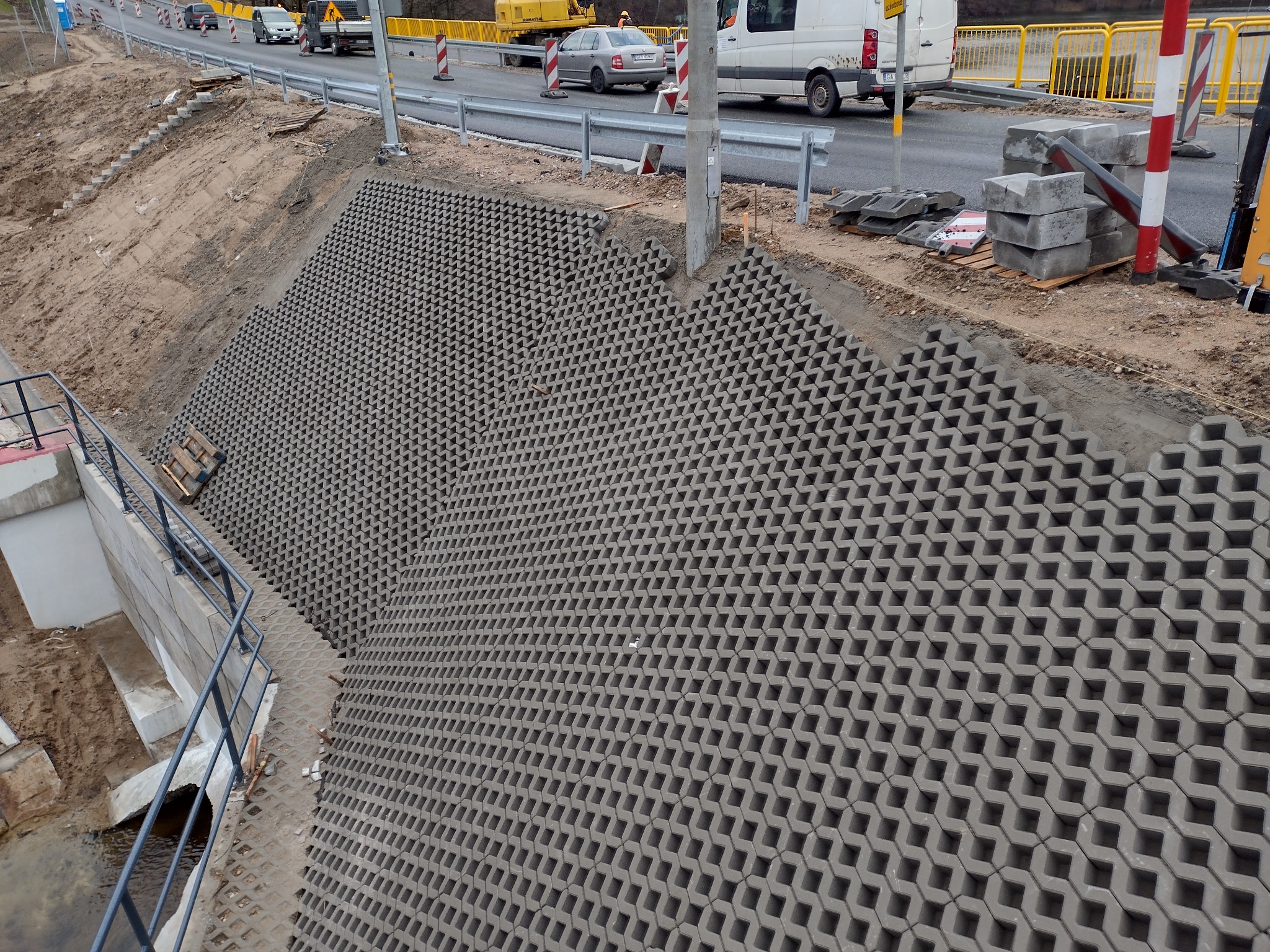 Wzmocnienie skarpy przy drodze za pomocą ażurowych płyt betonowych. Widoczne elementy infrastruktury drogowej i fragment przepustu wodnego.