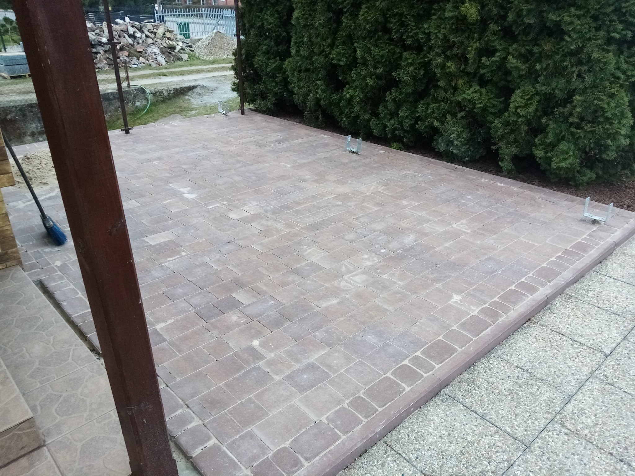 Ułożona kostka brukowa w odcieniach brązu, obrzeża z kostki, widoczne elementy konstrukcji zadaszenia, w tle tuje i materiały budowlane.