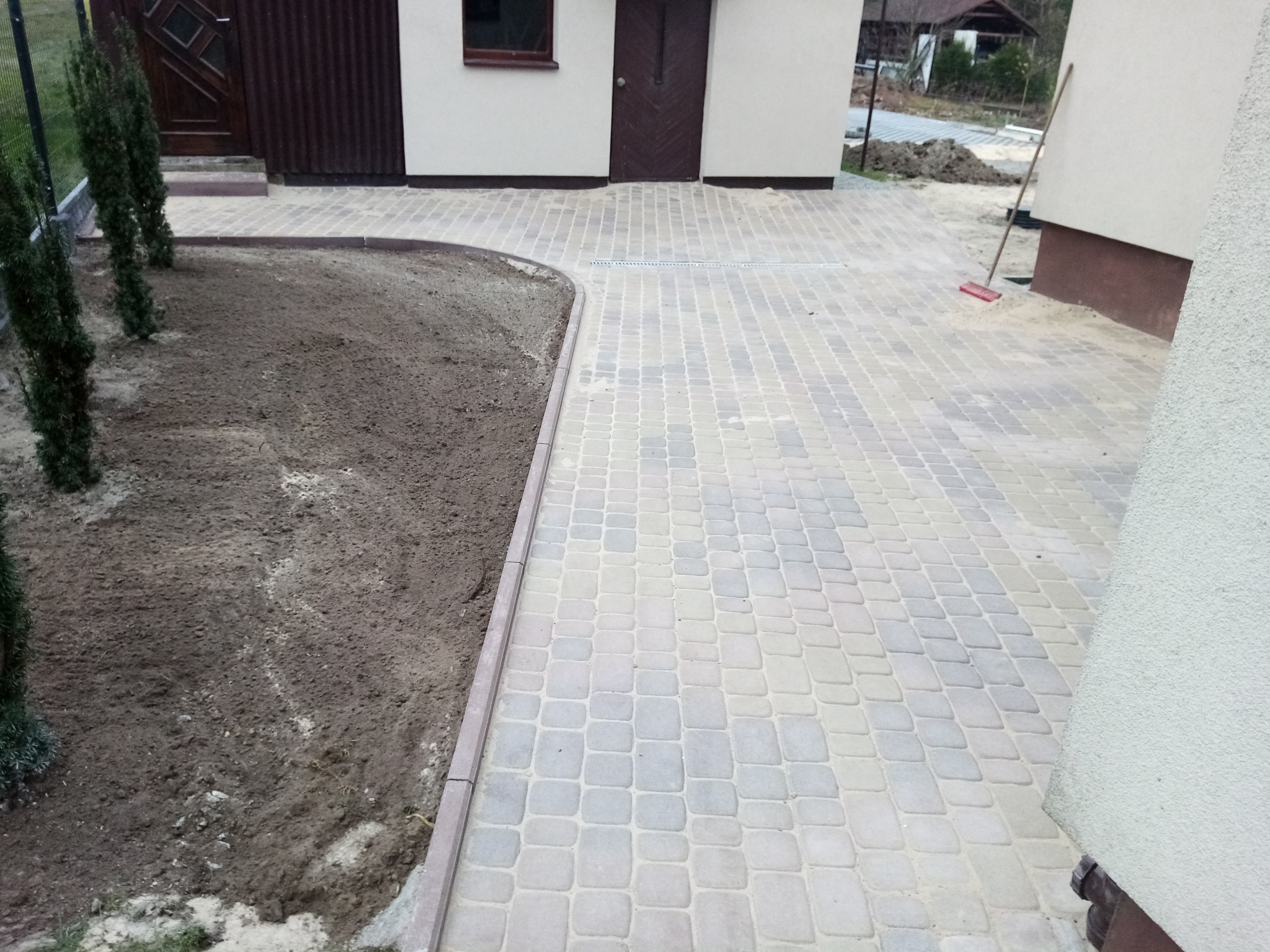 Ułożona kostka brukowa w odcieniach szarości i beżu tworzy ścieżkę obok przygotowanego terenu pod nasadzenia. Widoczny fragment budynku i narzędzie pracy.