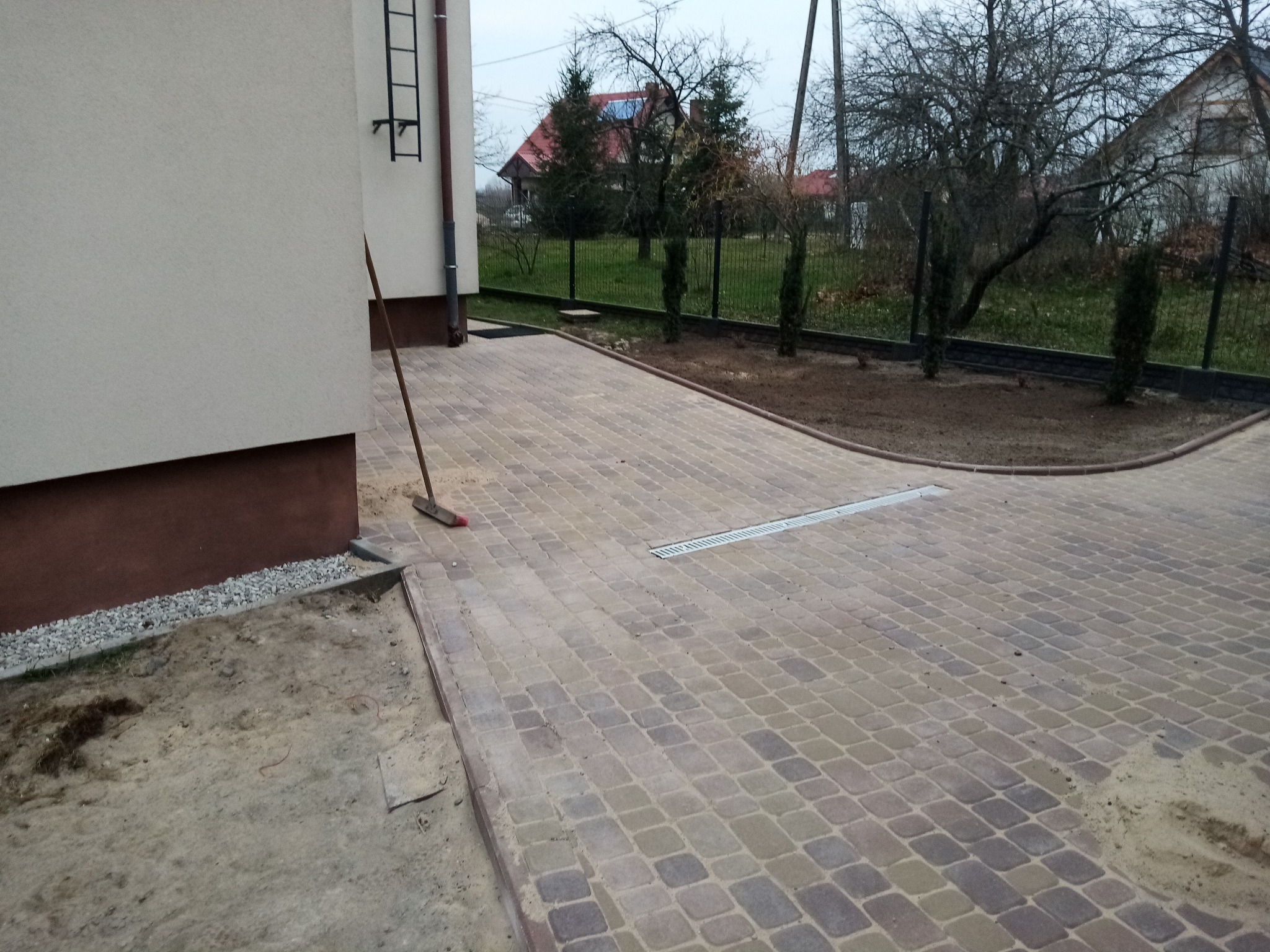 Ukończony podjazd z kostki brukowej w odcieniach beżu i szarości, z widocznym odpływem liniowym i fragmentem budynku z drabinką. W tle ogrodzenie panelowe i zieleń.