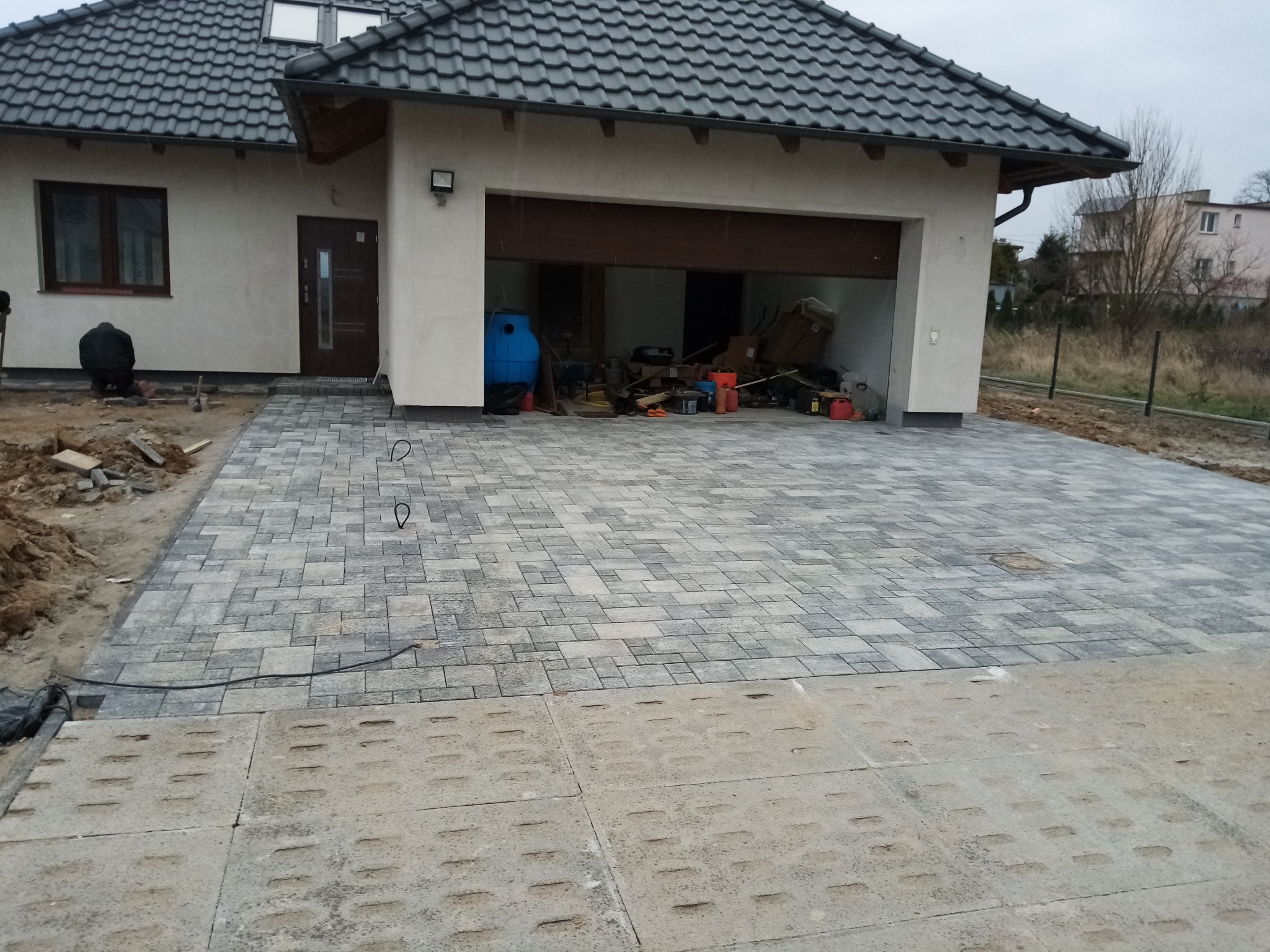 Ułożona kostka brukowa przed domem z otwartym garażem, widoczne prace wykończeniowe i przygotowanie terenu. Szara kostka w geometryczny wzór.