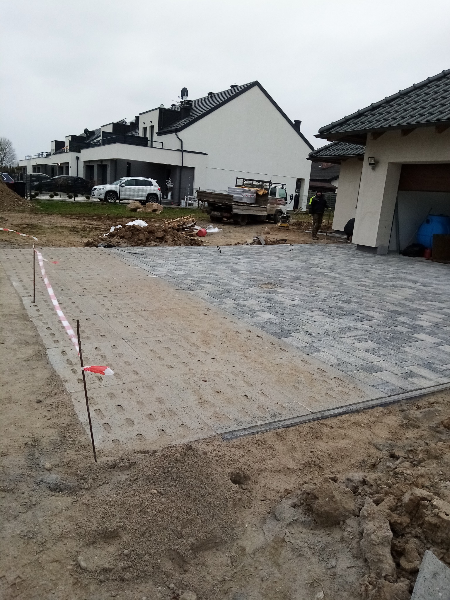 Częściowo ułożona kostka brukowa na podjeździe, widoczne etapy prac. W tle domy jednorodzinne i samochód dostawczy. Taśma ostrzegawcza wyznacza obszar robót.