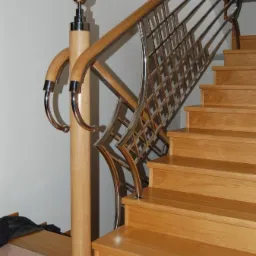 Schody z drewnianymi stopniami i balustradą ze stali nierdzewnej o ażurowym wzorze, zakończoną drewnianym poręczą. Widoczny detal słupka z ozdobnym zakończeniem.