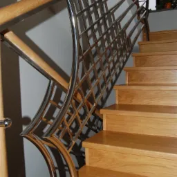 Schody z drewnianymi stopniami i unikalną, ażurową balustradą ze stali nierdzewnej, z drewnianym poręczem. Element dekoracyjny w tle, światło dzienne.
