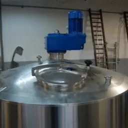 Błyszczący, metalowy zbiornik z niebieskim silnikiem na górze, otoczony drabinkami. Widoczny fragment hali produkcyjnej. Charakterystyczny okrągły właz.