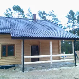 Drewniany dom z ciemną dachówką i rynnami, widok z zewnątrz. Fragment tarasu z drewnianą balustradą. W tle las iglasty.