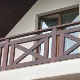 Drewniana balustrada balkonowa z krzyżującymi się elementami, widok z dołu, z fragmentem dachu i okna w tle. Ciemne drewno, jasne ściany.