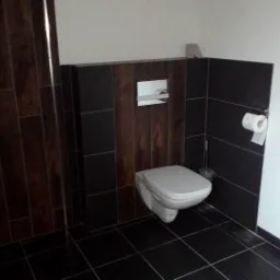 Nowoczesna toaleta z czarną glazurą imitującą drewno i białymi ścianami. Wisząca miska WC, chromowany przycisk spłukujący. Minimalistyczny design.