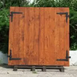 Rustykalna, drewniana ścianka działowa z ciemnymi okuciami na paletach, ustawiona na zewnątrz na tle zieleni. Widoczne naturalne przebarwienia drewna.