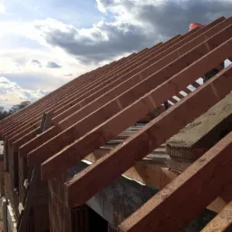 Konstrukcja dachu w trakcie budowy z widocznymi krokwiami, murłatami i pustakami. W tle błękitne niebo z chmurami i fragment zabudowy mieszkalnej.