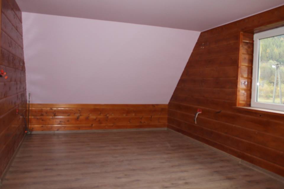 Pokój na poddaszu z drewnianą boazerią do połowy ściany i panelami na podłodze. Widoczne okno z widokiem na drzewa.