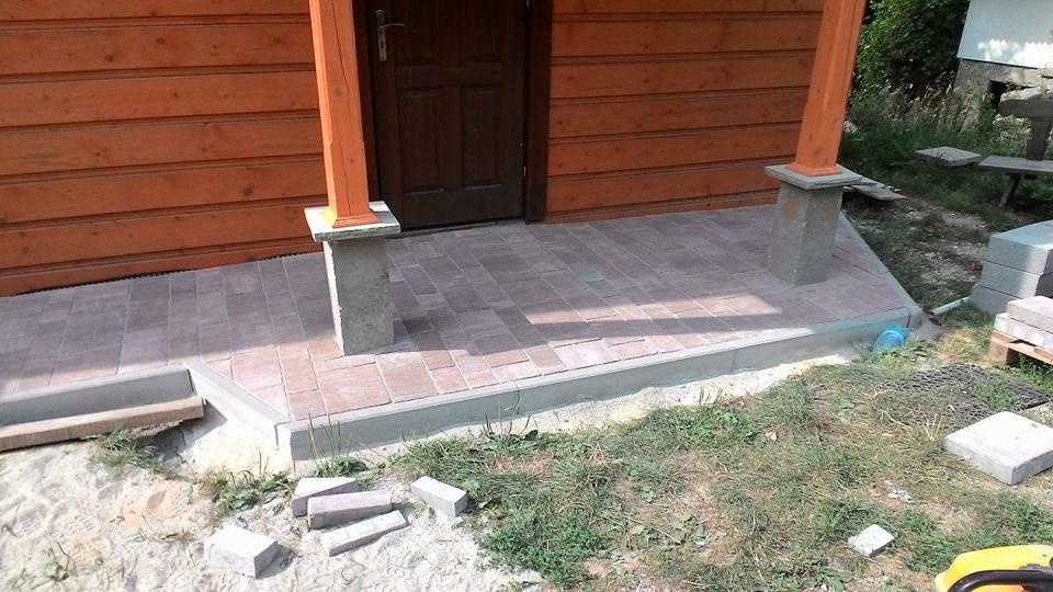 Wykończone wejście do drewnianego domu z brązową kostką brukową, betonowymi obrzeżami i filarami. Widoczne detale wykończenia i elementy otoczenia, takie jak trawa i niezabudowane kostki.