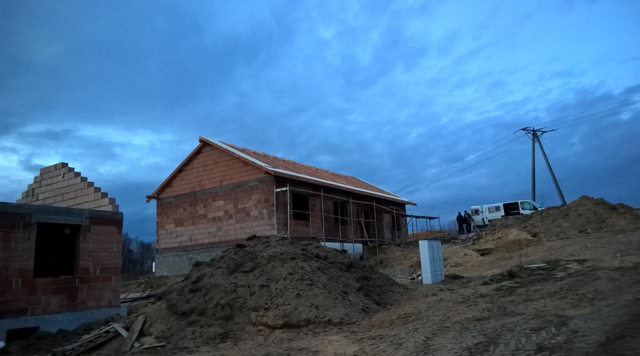 Wznoszenie domu z cegły: stan surowy otwarty, widoczne rusztowania i częściowo ułożony dach z dachówki. Na drugim planie ekipa budowlana i samochód dostawczy.