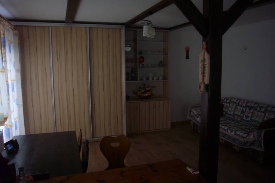 Jasne wnętrze pokoju z drewnianą szafą przesuwną, przeszkloną witryną i drewnianym stołem z krzesłami. Widoczny fragment sofy i drewniane elementy konstrukcyjne sufitu.