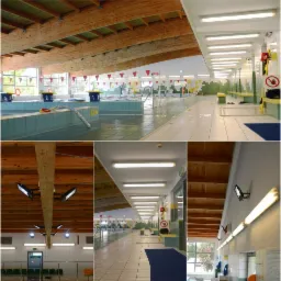 Oświetlony basen kryty z drewnianą konstrukcją dachu, lampy liniowe i reflektory. Widok na pusty basen i oświetlony korytarz z jasną, ceramiczną posadzką.