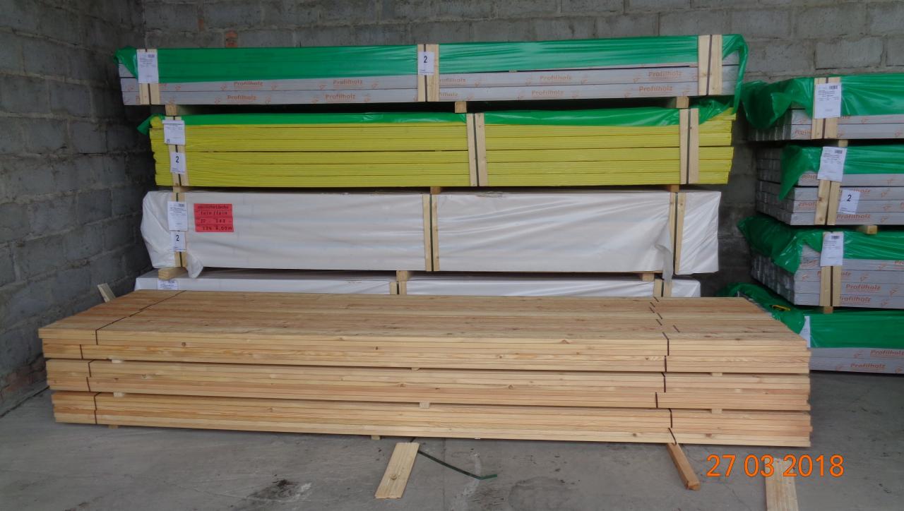 Stos tarcicy budowlanej ułożony w magazynie, widoczne pakiety w folii o różnych kolorach (zielony, żółty, biały) oraz pojedyncze deski na podłodze, data 27.03.2018 w prawym dolnym rogu.