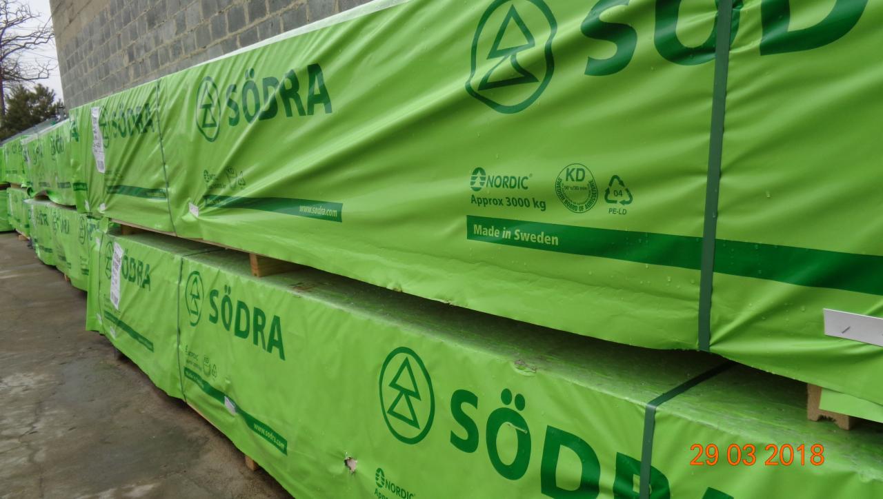 Stosy tarcicy budowlanej marki Södra na drewnianych paletach, zabezpieczone zieloną folią z nadrukowanymi informacjami o produkcie, w tym wadze i miejscu produkcji, składowane na zewnątrz przy...