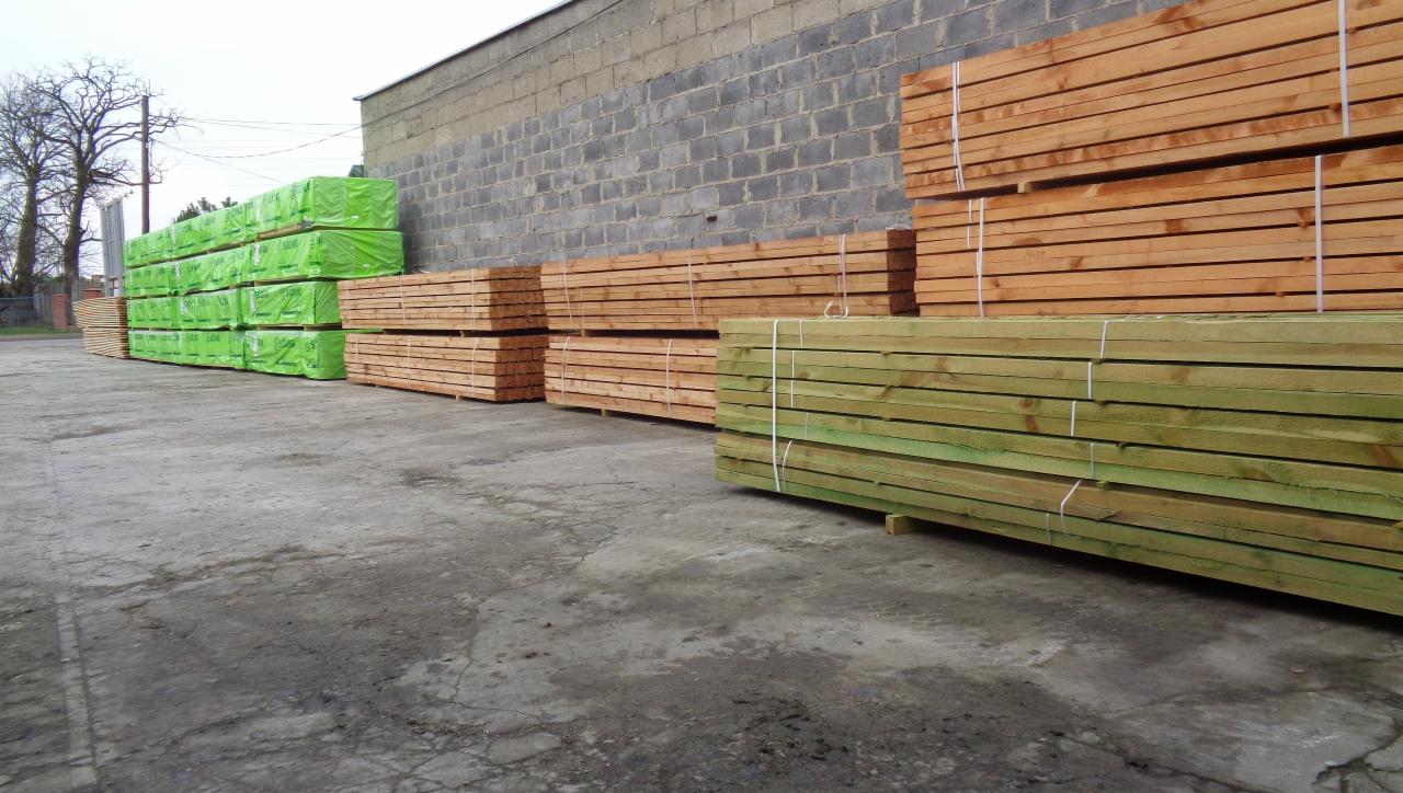 Stosy tarcicy budowlanej, część zafoliowana na zielono, ułożone na betonowym placu przy szarym, ceglanym budynku.