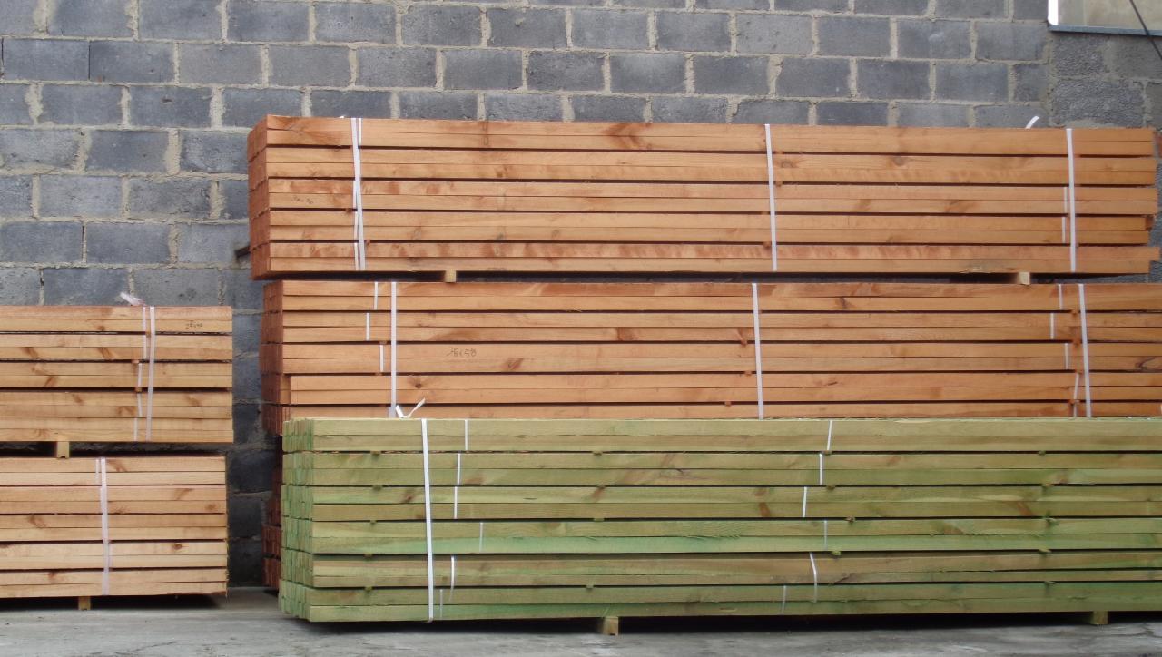 Ułożone w stosy paczki tarcicy budowlanej w różnym kolorze drewna, zabezpieczone białym sznurkiem, na tle szarej ściany z betonowych pustaków.