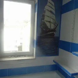 Wykończona łazienka z biało-niebieskimi płytkami i dekoracyjnym panelem z motywem żaglowca, widoczna wanna i okno z parapetem.