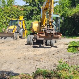 Dariusz Ryś - Żółta koparka kołowa i koparko-ładowarka na terenie budowy, widoczne ślady prac ziemnych i wycinki, w tle drzewa iglaste i liściaste pod błękitnym niebem.