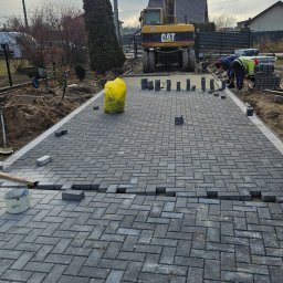 Dariusz Ryś - Montaż szarej kostki brukowej w jodełkę na posesji, z widoczną koparką CAT w tle i pracownikami układającymi obrzeża.